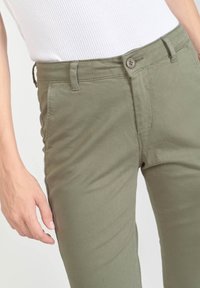 Olijfgroen, smal gesneden katoenbroek met voorzakken, een knoopsluiting en een gladde textuur. Zichtbare stiksels langs de naden.