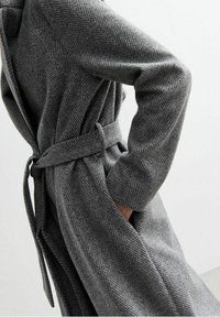 Abrigo gris con patrón de espiga, de mangas largas, con un cinturón atado en la cintura y una tela texturizada, que muestra un ajuste entallado y detalles sutiles.