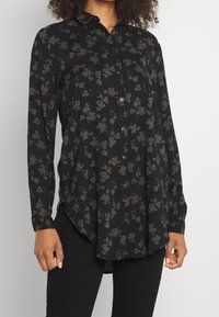 Camisa negra de botones con manga larga, que presenta un patrón floral en verde oscuro. Tejido suave con un dobladillo curvado y un brillo sutil.
