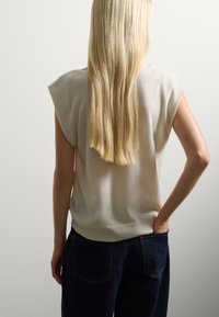 Beige ärmelloser Pullover mit geripptem Saum, weicher Textur und lockerer Passform, kombiniert mit dunkelblauen hochtaillierten Jeans. Glatter Rückansicht.