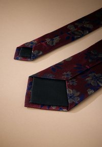 Cravate en tissu bordeaux avec des motifs floraux en or et bleu, présentant une extrémité pointue avec un dessous noir lisse. Surface texturée.
