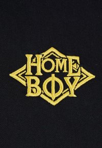 Črni tekstilni aplikator z izvezenim besedilom "HOME BOY" v rumeni barvi, s dekorativnim diamantnim obriscem in ostrimi poudarki.