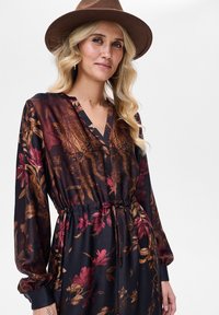 Długa sukienka w kwiaty z ciemnego materiału, z dekoltem w serek, wiązaniem w talii oraz długimi, luźnymi rękawami. Wzór kwiatowy w kolorach czerwonym i złotym.