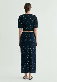 Mujer con el pelo recogido que lleva un conjunto de top corto azul marino y falda larga con estampados blancos de cachemira y flores, de pie descalza sobre un suelo claro.