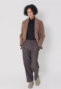 Brun krävd kappa över en svart höghalsskjorta, ihop med grå rutiga högmidjade byxor. Skorna är ljusbruna mocka ankelstövlar.