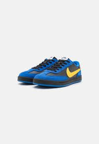 Kék velúr sportcipők fekete bőr részletekkel és sárga Nike logóval. Fekete fűzőkkel és minimális barázdázottságú gumitalppal rendelkezik.