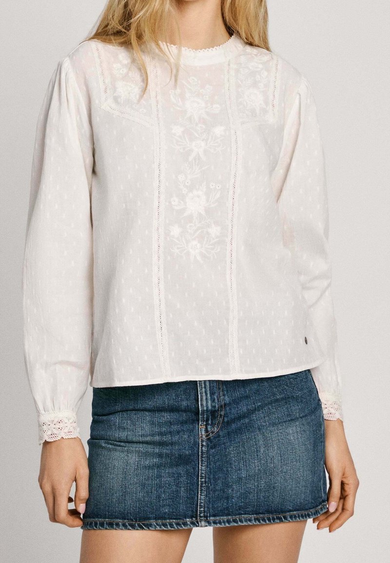 Frau, die eine weiße Bluse mit langen Ärmeln und floralen Stickereien sowie Spitzenakzenten trägt, kombiniert mit einem blauen Jeans-Minirock.