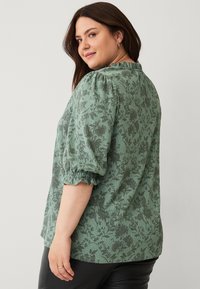 Grøn blomstret bluse med pufærmer og flæsekanter. Har et struktureret stof med mørkere grønne blomsterprint. Vist fra ryggen.