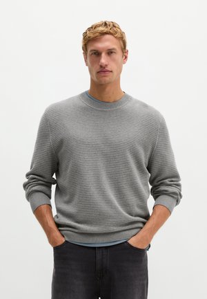Mann mit blondem Haar, der einen hellgrauen, gerippten Pullover und dunkle Hosen trägt, steht mit den Händen in den Taschen vor einem weißen Hintergrund.