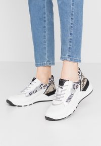 Bílé chunky tenisky s kombinovanými zvířecími potisky, obuté s světle modrými džíny, na prosté bílé podlaze a zdi.