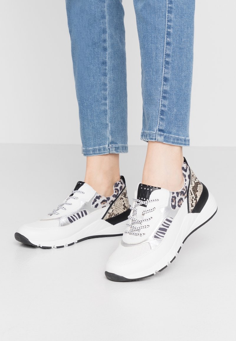 Bílé chunky tenisky s kombinovanými zvířecími potisky, obuté s světle modrými džíny, na prosté bílé podlaze a zdi.