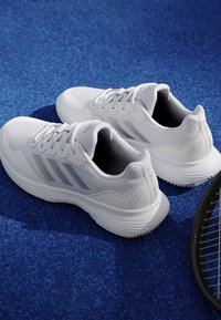 White athletic shoes with gray stripes on a blue textured surface next to a black tennis racket.