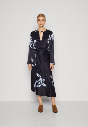 Satin wrap dress in navy with white floral patterns, deep V-neck, long sleeves, tied waist, and a flowing hemline. Brown open-toed shoes.