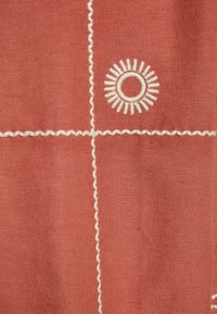 Il tessuto terracotta presenta un design a sole ricamato in bianco con cuciture a zig zag, creando un motivo contrastante su una texture liscia.