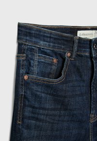Jeans de mezclilla azul oscuro con una textura suave, diseño de cinco bolsillos, con herrajes de botón de cobre y costuras doradas en contraste.