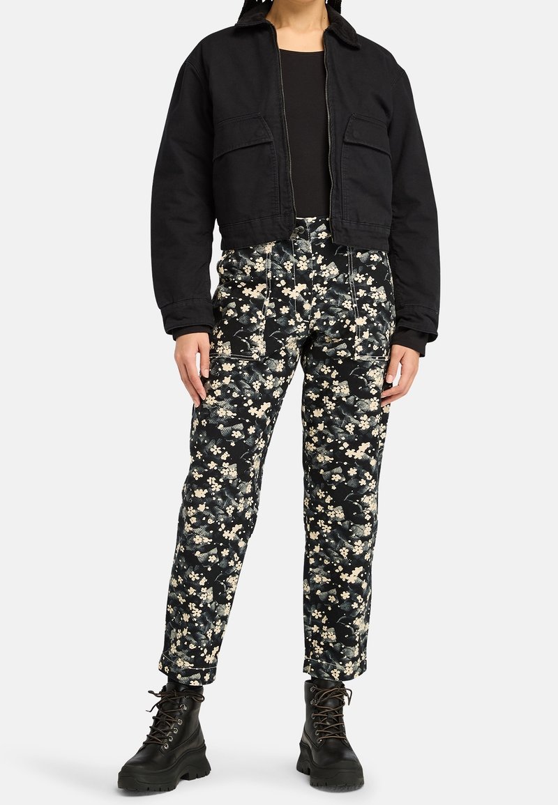 Personne portant une veste courte noire, un haut noir, un pantalon noir à motifs floraux et des bottes noires à lacets, debout devant un fond blanc.