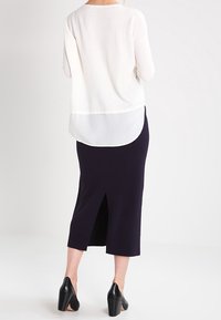 Top de malla blanca superpuesto sobre una base blanca transparente, combinado con una falda lápiz azul marino con una abertura en la parte trasera. Zapatos de tacón bloque negro completan el look.