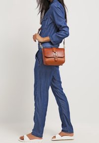 Hnědá kabelka z umělé kůže s texturovaným povrchem, obdélníkového tvaru, a zlatými kovovými detaily, nošená přes tělo s džíny.