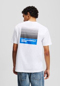 Balta medvilninė marškinėliai su horizontaliomis juodomis ir mėlynomis juostelėmis, o nugaros pusėje yra užrašas "KARL LAGERFELD JEANS". Laisvas kirpimas ir trumpomis rankovėmis.