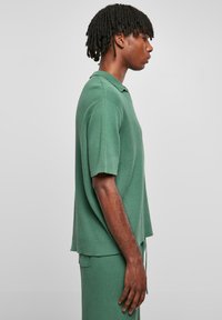 Grønn strikket polo-skjorte med korte ermer, krage og løs passform. Det teksturerte stoffet har vertikale ribbestrikkede detaljer.