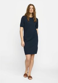 Robe tricotée bleu marine à manches courtes, encolure ronde et fentes sur les côtés. Portée avec des sandales tressées marron. Tissu lisse de poids moyen.