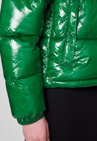 Casaco puffer verde brilhante com um design acolchoado, punhos elásticos e um bolso no peito, combinado com uma peça preta canelada abaixo.
