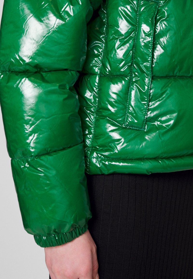 Glanzende groene pufferjack met een gewatteerd ontwerp, elastische manchetten en een borstzak, gecombineerd met een geribbelde zwarte kleding onderaan.
