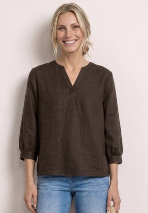 Uśmiechnięta kobieta z blond włosami upiętymi w niski kok nosi ciemnobrązową bluzkę z długim rękawem i jasnoniebieskie dżinsy, trzymając jasny przedmiot w prawej ręce.