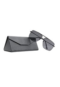 Occhiali da sole con montatura geometrica nera appoggiati su un astuccio pieghevole triangolare nero con texture, su uno sfondo bianco.