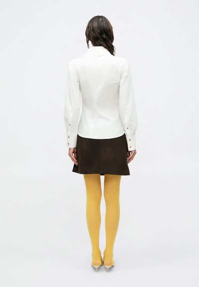 Biela blúzka s dlhým rukávom a priliehavým strihom, hnedá mini sukňa zo semišu, žlté pančuchy a strieborné lodičky so špicatou špičkou. Jednoduchý a moderný vzhľad.