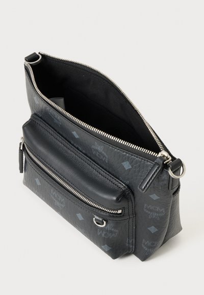 Μαύρη MCM τσάντα crossbody με ασημένια φερμουάρ, μπροστινή τσέπη, λεπτομέρεια δαχτυλιδιού και μοτίβο μονόγραμμα, εμφανίζεται ανοιχτή σε ανοιχτό φόντο.