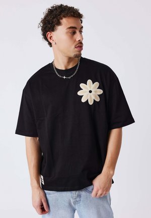 Jonge man met krullend haar, gekleed in een zwart T-shirt met een groot beige bloemmotief, lichtblauwe spijkerbroek en een zilveren kettinkje.