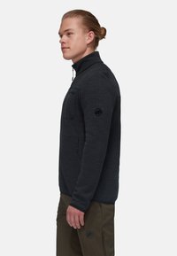 Giacca in pile grigio scuro con colletto alto, tasca sul petto con zip e logo nero sulla manica, abbinata a pantaloni marrone chiaro.