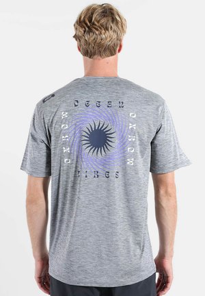 Homme aux cheveux blonds courts portant un T-shirt gris clair avec un motif en spirale de soleil violet et le texte « OCEAN VIBES » au dos.