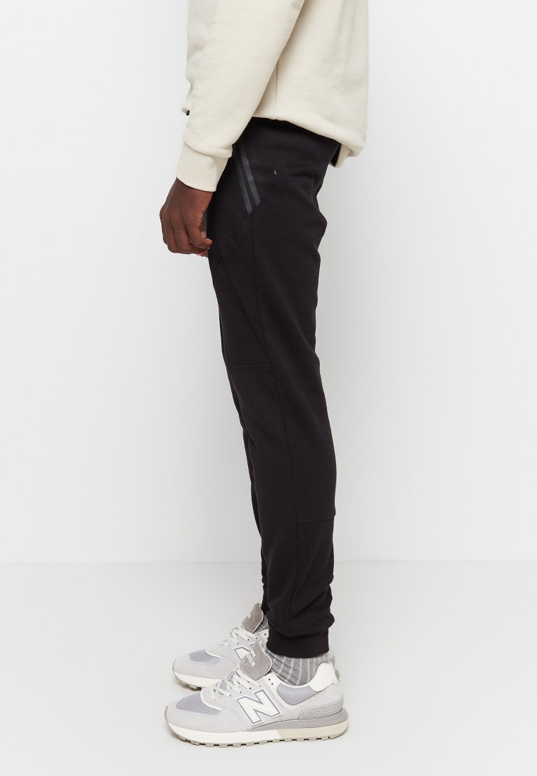 Los joggers negros ajustados cuentan con una cintura elástica, bolsillos laterales y detalles de franjas en contraste. Se combinan con zapatillas deportivas grises.