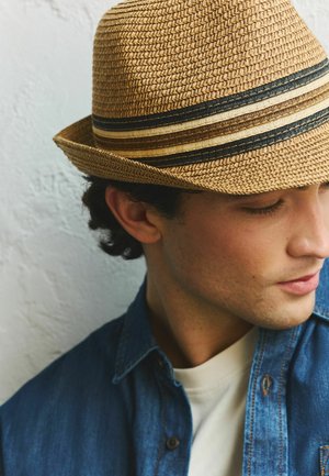 Ung mand iført en flettet stråhat med sorte, brune og beige striber, der kigger ned, iført denimjakke og hvid skjorte.