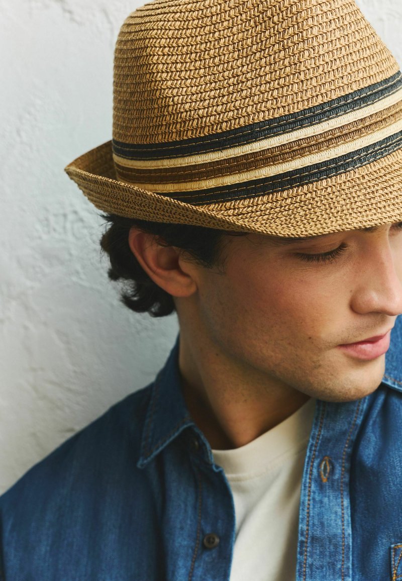 Fiatal férfi, aki szalmaszövésű, fekete, barna és bézs csíkos fedora kalapot visel, lefelé néz, farmerdzsekit és fehér inget visel.
