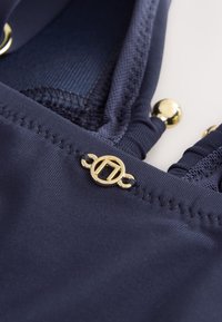 Gros plan sur un tissu bleu marine avec un logo circulaire doré et des perles dorées décoratives attachées à des bretelles froncées.