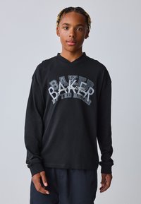 Sort langærmet T-shirt med V-udskæring, prydet med hvid og grå grafisk tekst "BAKER" på forsiden. Stoffet virker glat og afslappet.