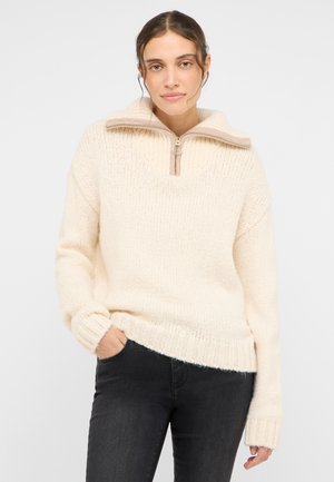 Cremefarbener Pullover mit einem halben Reißverschluss, gerippten Bündchen und Saum sowie einem weichen, strukturierten Strick. Kombiniert mit schwarzen Jeans.