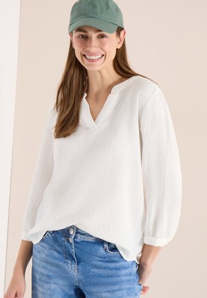 Lächelnde Frau mit grüner Mütze, weißer, strukturierter Langarmbluse mit V-Ausschnitt und blauen Jeans vor beigem Hintergrund.