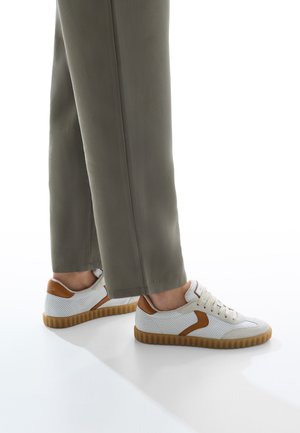 Person iført olivengrønne bukser og hvide sneakers med brune detaljer og gummisåler, stående på en hvid reflekterende overflade.