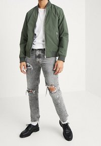 Man som bär en olivgrön bomberjacka, vit t-shirt, slitna grå jeans, vita strumpor och svarta snörskor, stående mot en vit bakgrund.