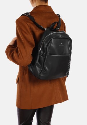 Zaino in pelle nera con un design arrotondato, dotato di tasca frontale con cerniera e dettagli testurizzati. Indossato sopra un cappotto in suede marrone.