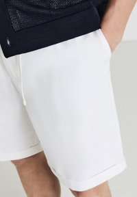 Person, der weiße Shorts mit Taschen trägt, die Hand im rechten Taschen, kombiniert mit einem navyblauen Strickoberteil mit Knopfdetaillierung.