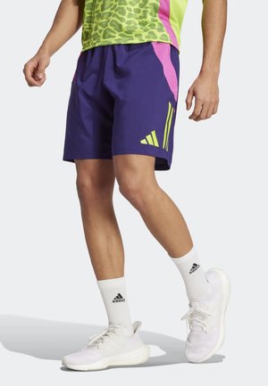 Mann trägt lila Sportshorts mit neongelben und pinken Akzenten, weiße Adidas-Socken und weiße Sportschuhe, steht auf grauem Boden.