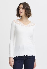 Weißes Langarmoberteil mit gewelltem Saum und Spitzen-V-Ausschnitt, mit gerippter Textur und tailliertem Design. Getragen mit dunkelblauen Jeans.