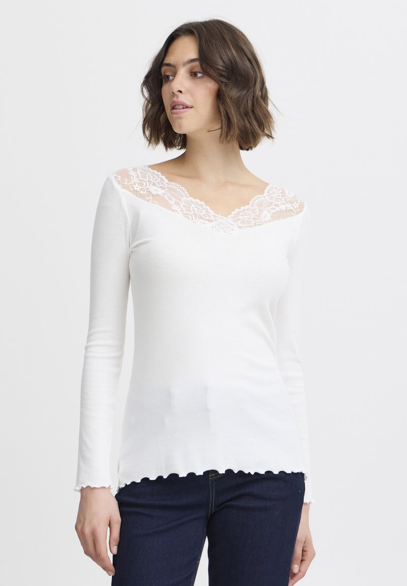 Weißes Langarmoberteil mit gewelltem Saum und Spitzen-V-Ausschnitt, mit gerippter Textur und tailliertem Design. Getragen mit dunkelblauen Jeans.