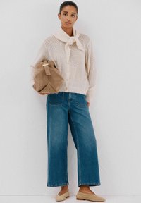 Cardigan in crema lavorato a maglia con colletto e fiocco, abbinato a jeans in denim blu a gamba larga e mocassini in suede beige, mentre tiene una borsa marrone.