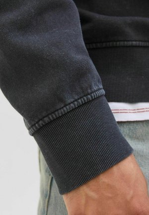 Schwarzer Sweatshirtbund mit geripptem Stoff und Nähdetails. Sichtbare Schichtung mit einem weißen Hemd darunter und einem Jeans-Hintergrund.